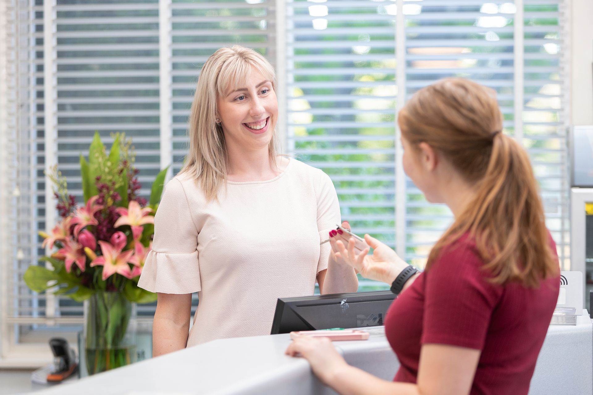 Two women at a reception desk exchanging a key, with flowers and blinds in the background