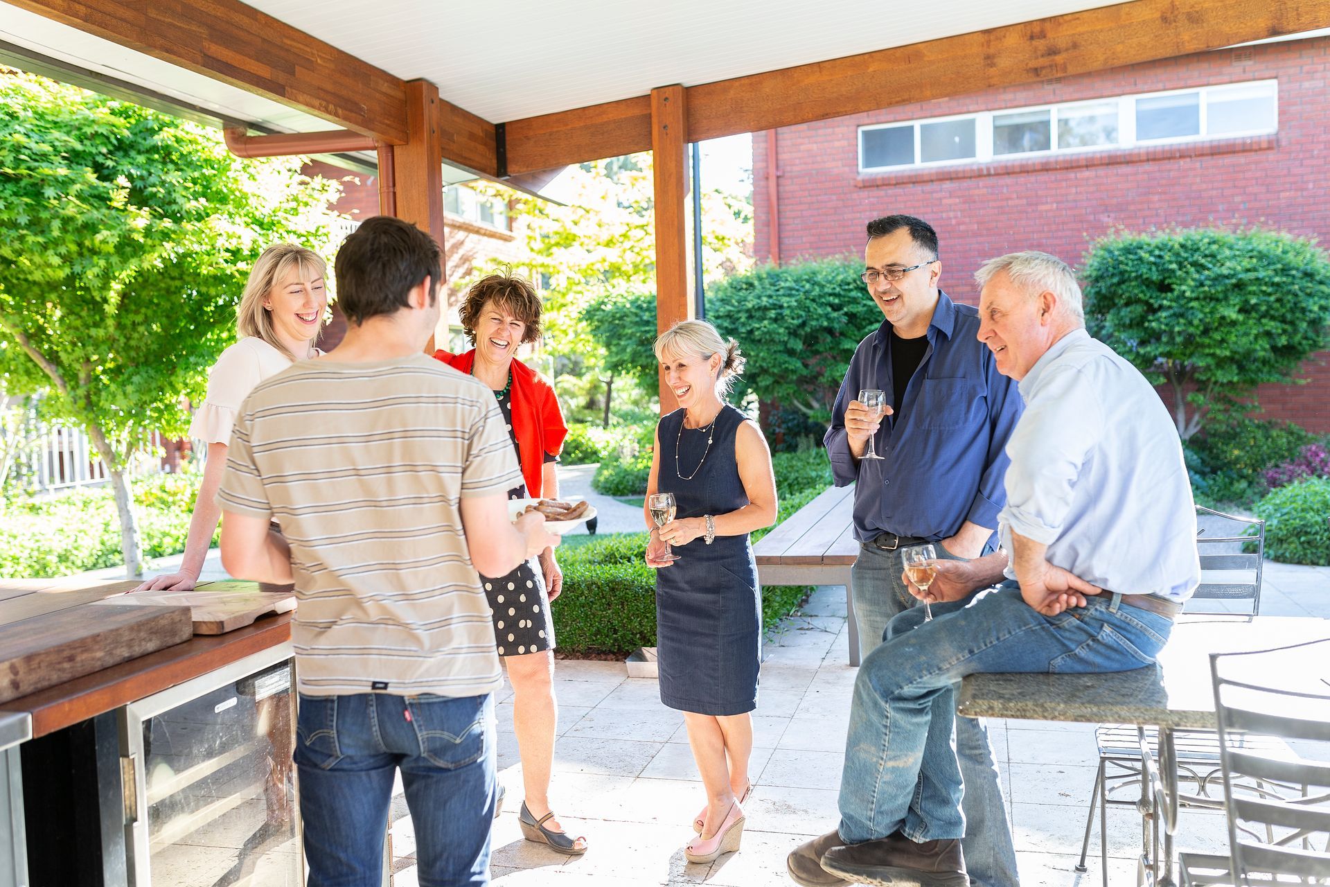 People chatting under a covered patio beside a brick building, with one man sitting on a stool