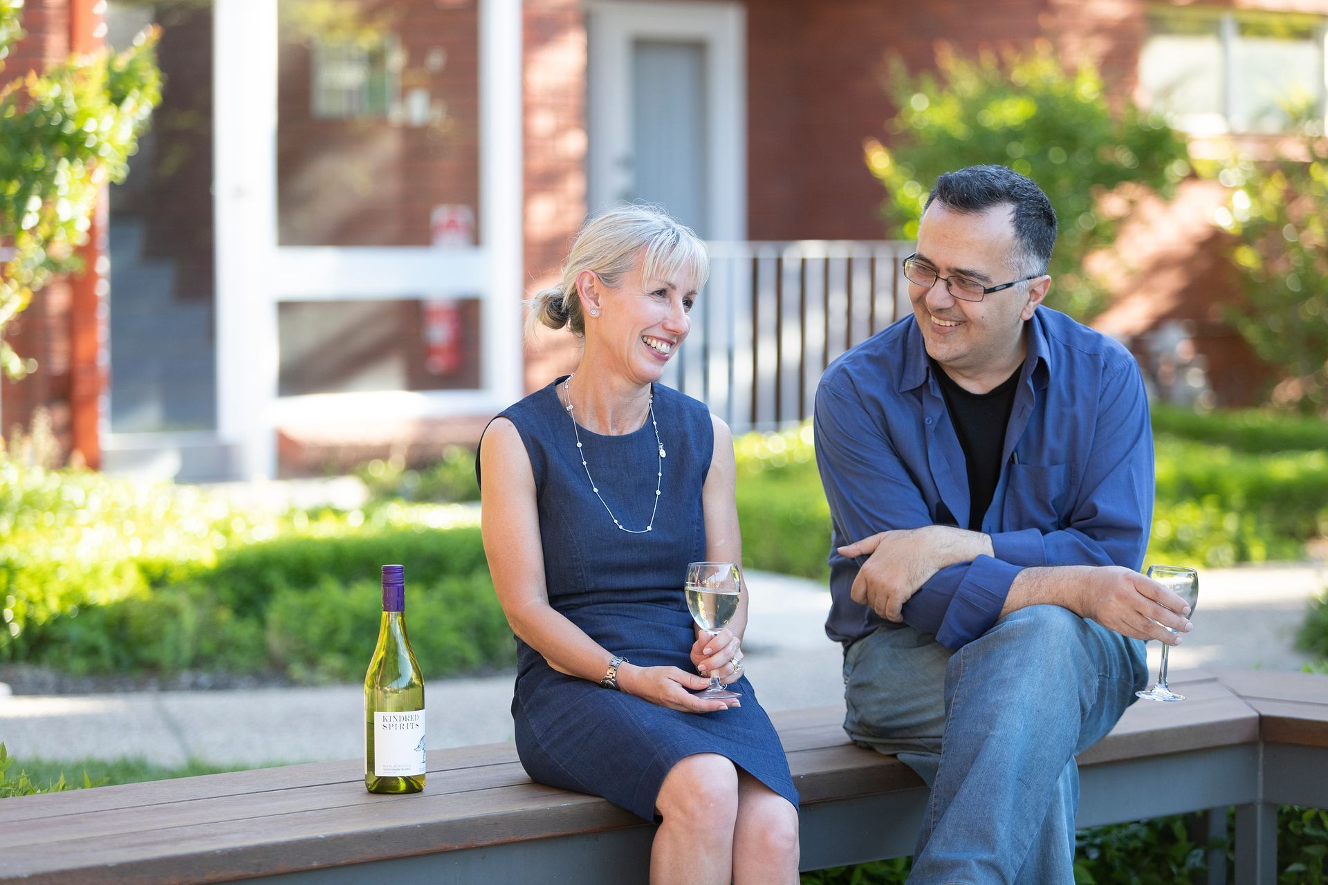 Two people sitting on an outdoor bench, smiling and chatting with drinks beside a house.
