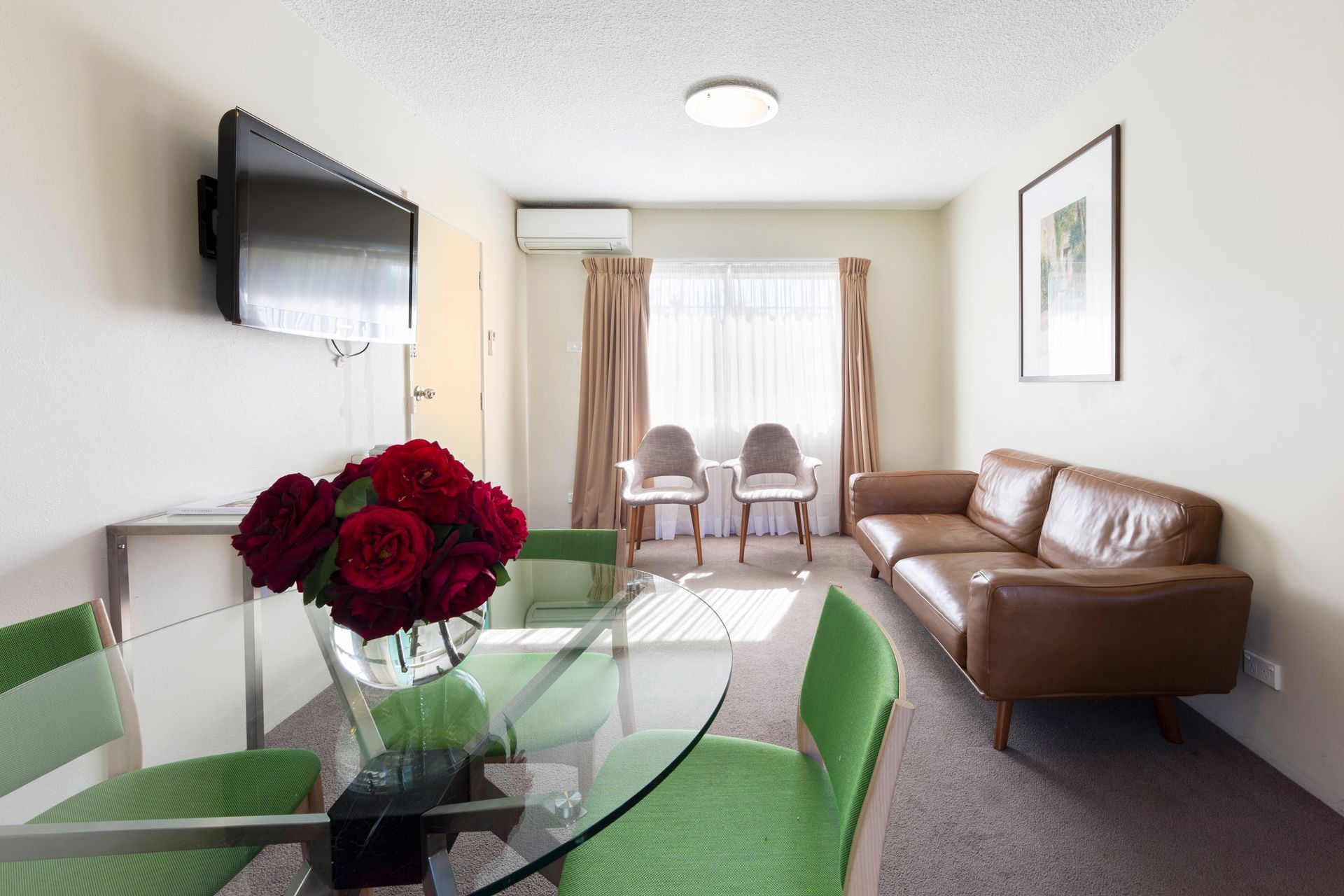 Bright living room with brown sofa, glass table, green chairs, wall TV, and red flowers by a window.