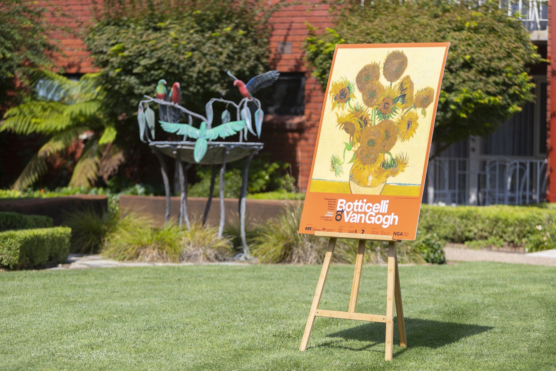 Lawn display with a painted easel sign and a turquoise sculpture in front of a brick building.