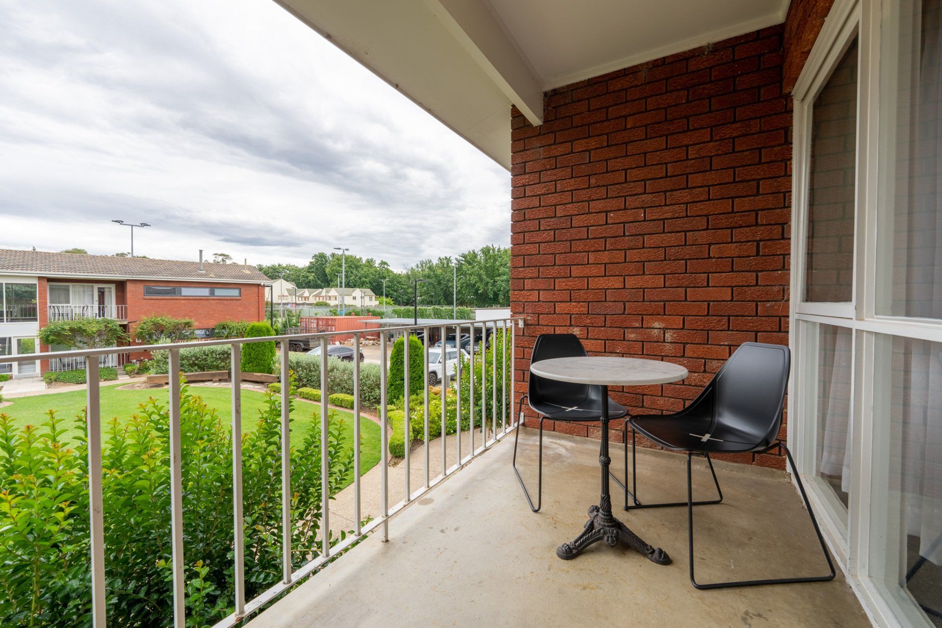 Balcony with black chairs and table beside a brick wall, overlooking a green courtyard and apartment buildings.