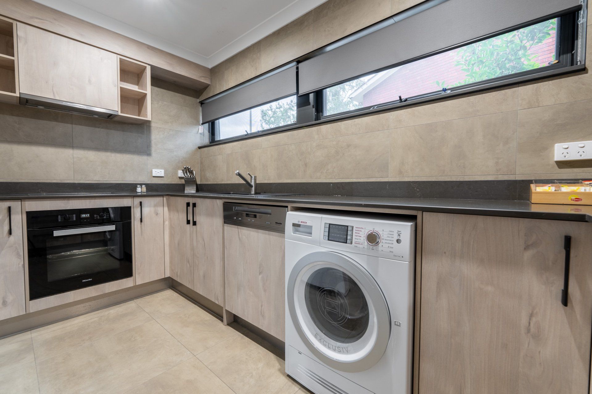 Modern kitchen with light wood cabinets, black countertops, built-in oven, washer, and long narrow windows
