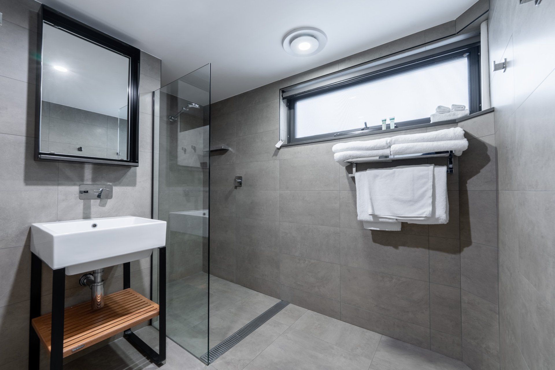 Modern bathroom with gray tile, glass shower, sink, mirror, and towel rack by a window.