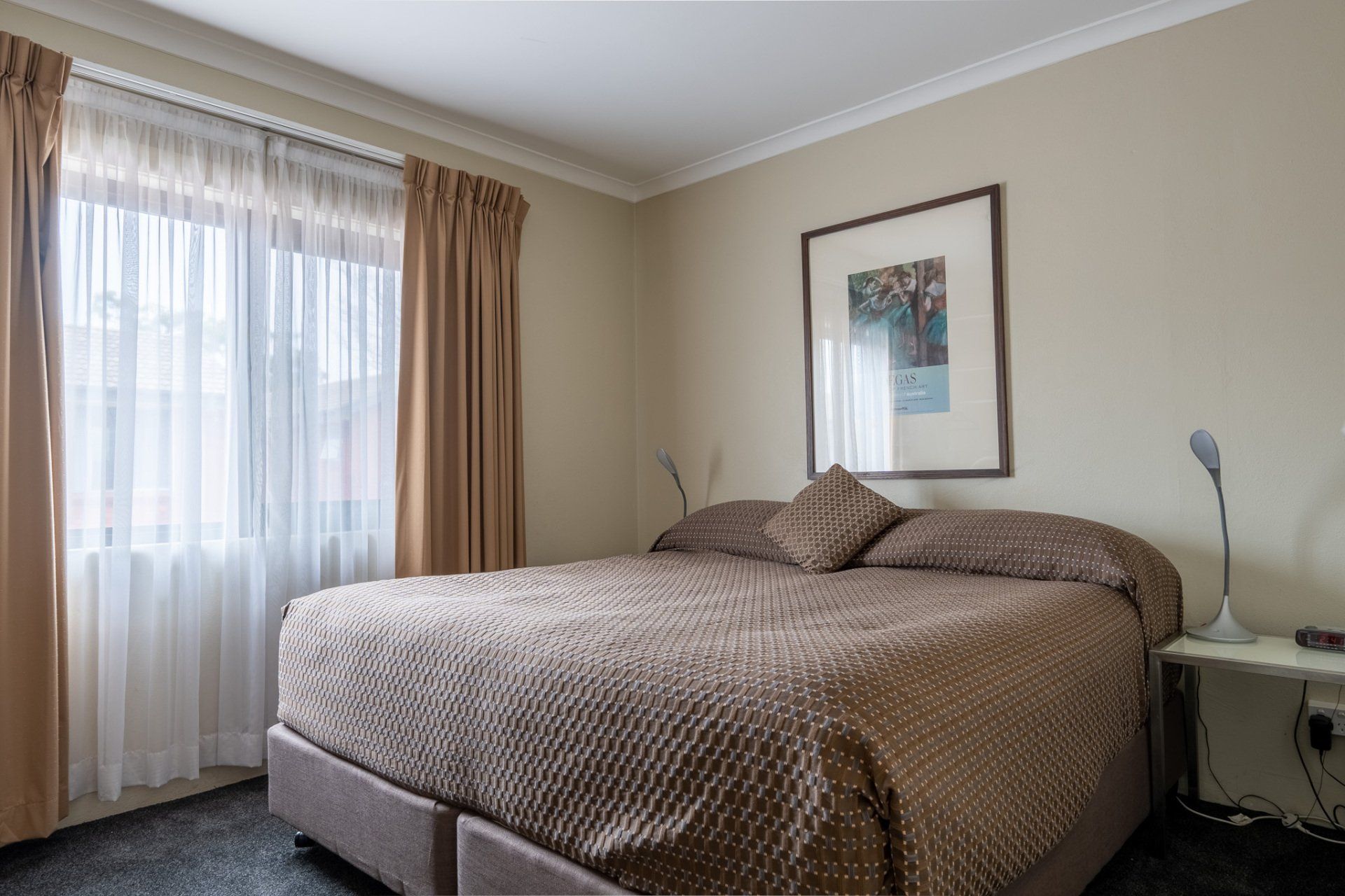 Bedroom with a large bed, bedside table, framed art, and a window with sheer curtains.