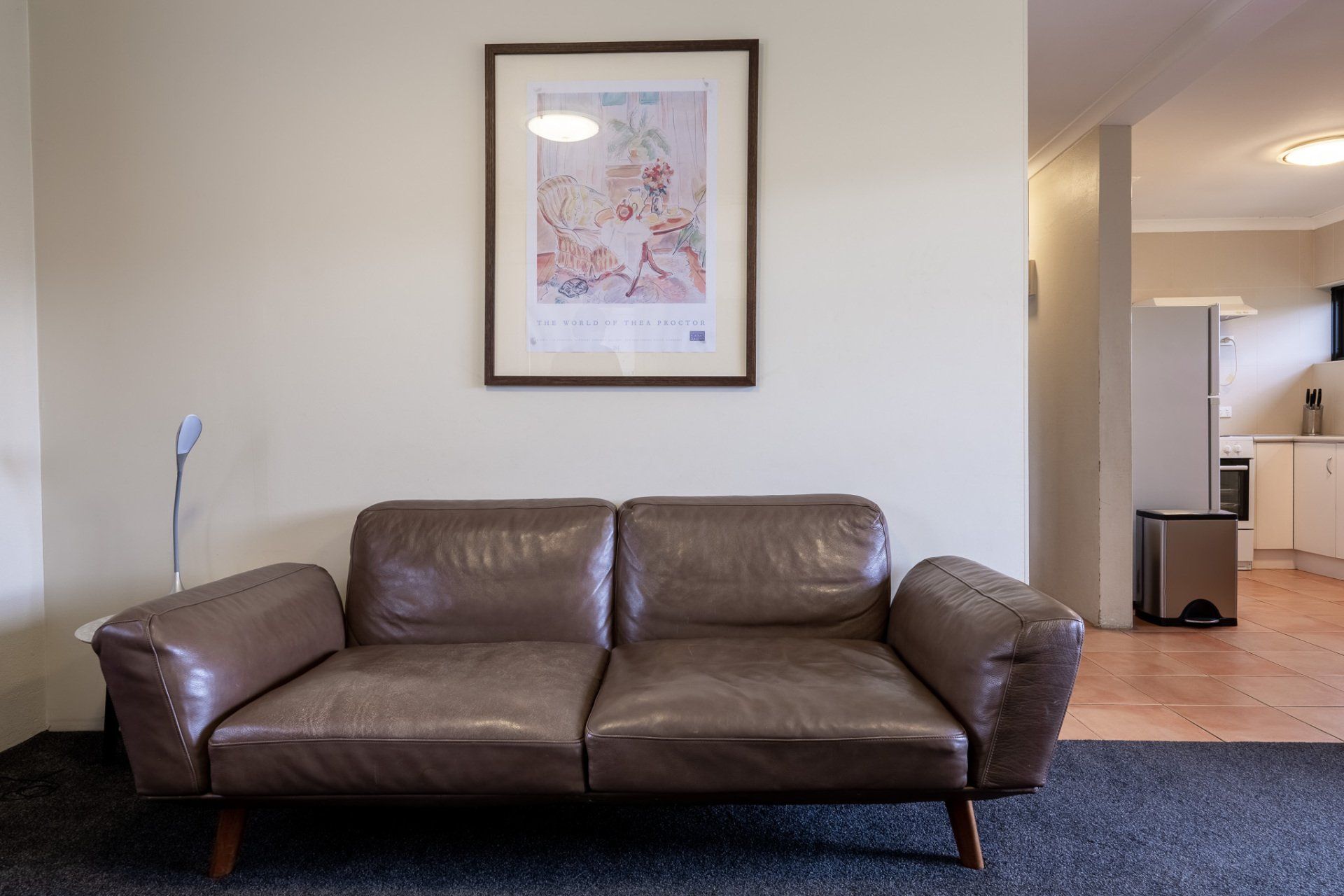 Brown leather sofa against a beige wall with framed art, in a living room opening to a kitchen.