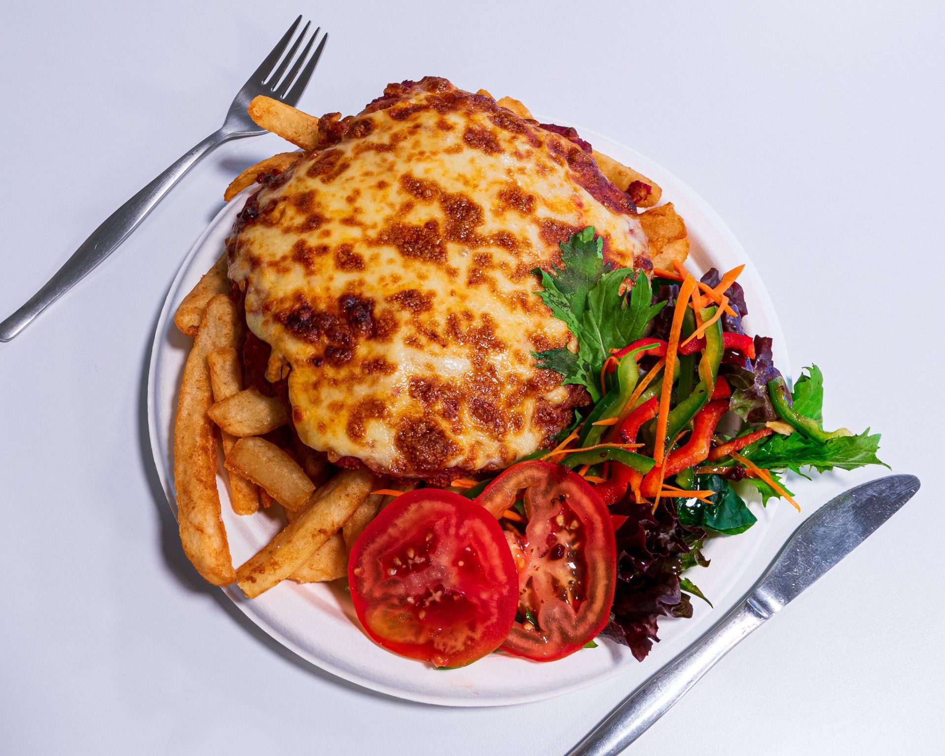 Baked cheesy pasta on a plate with fries, tomatoes, greens, and a fork and knife