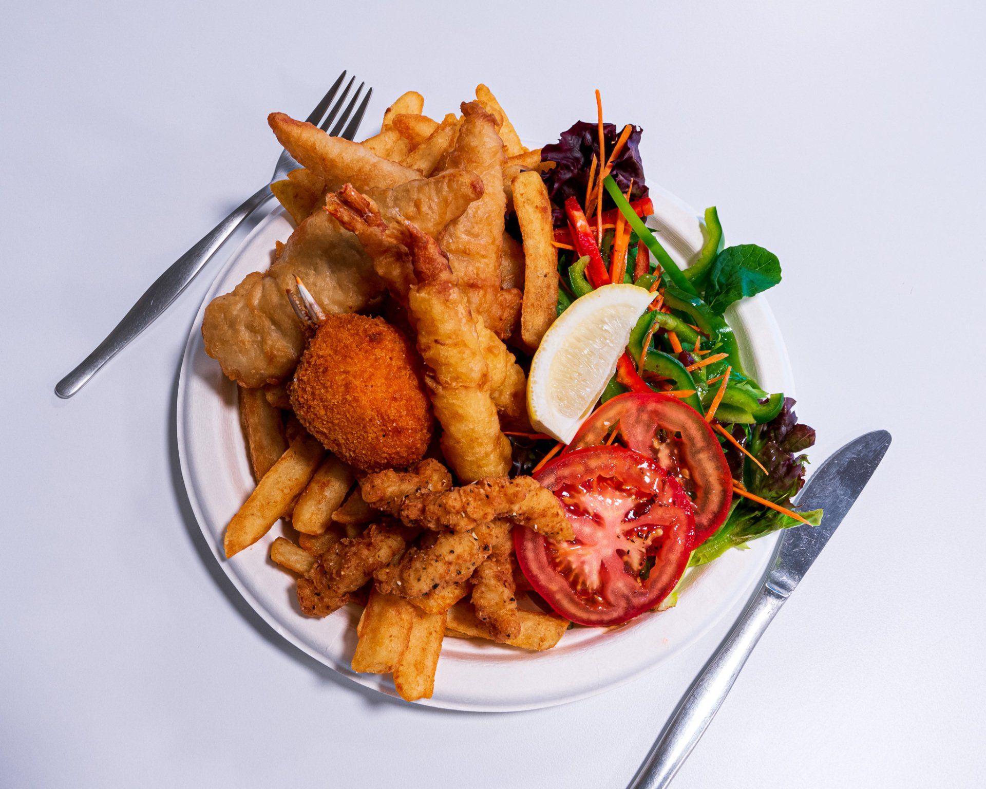 Plate of assorted fried seafood and fries with salad and lemon wedges on a white table