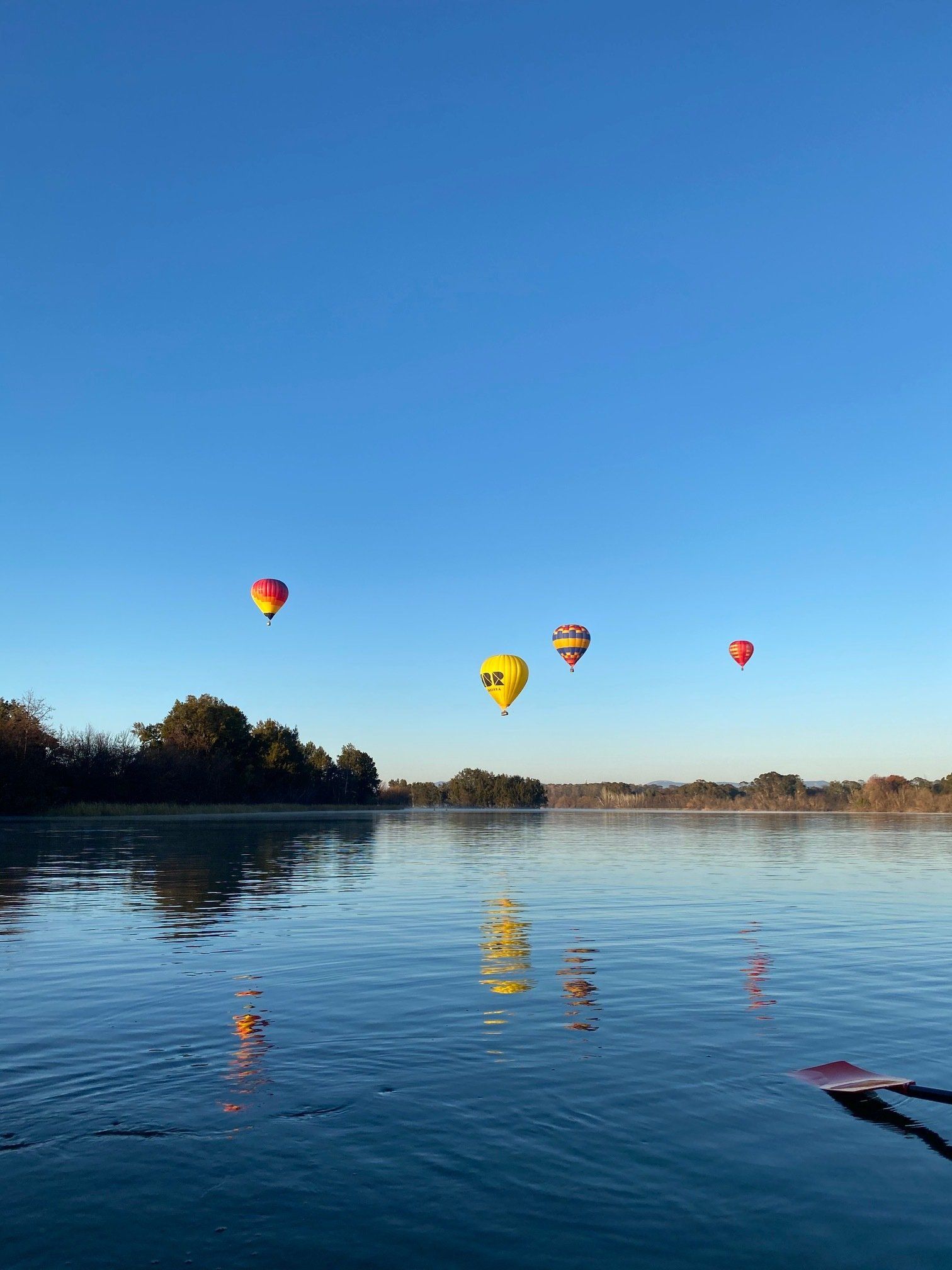 Colorful hot air balloons float above a calm river at sunrise, with reflections on the water.