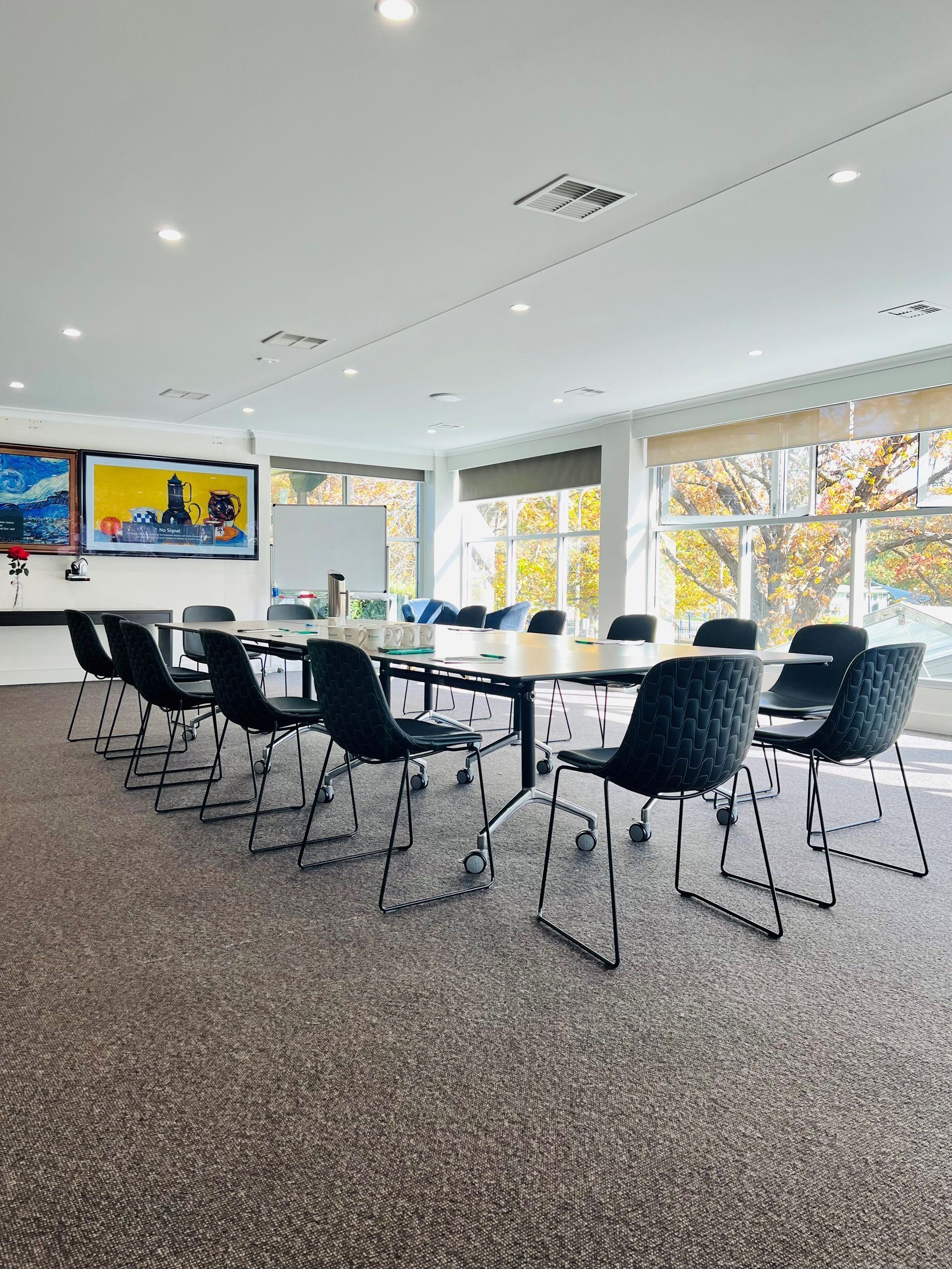 Modern conference room with long table, black chairs, wall art, and large windows overlooking autumn trees