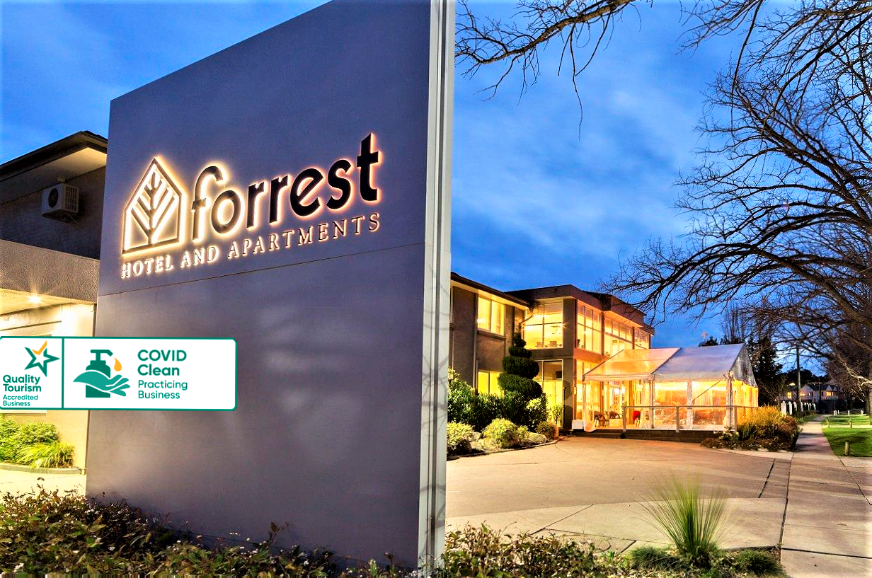 Evening exterior of Forrest hotel and apartments, with lit sign, parking lot, and illuminated building.