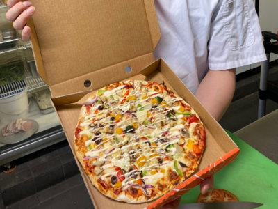 Open pizza box with a topped pizza on a person’s lap at a green table