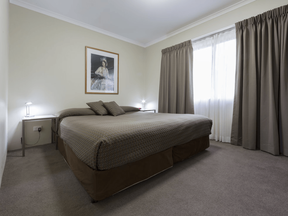 Neutral-toned bedroom with a made bed, nightstands, framed art, and curtains over a bright window