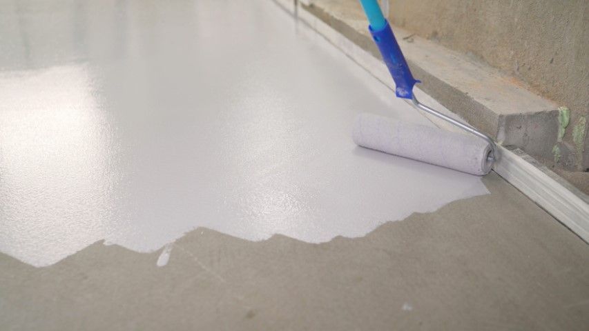 Blue-handled paint roller applying white coating to a floor edge beside a wall.