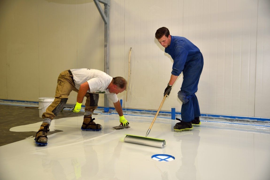 Two workers smoothing a white floor with a long-handled squeegee in a room under renovation