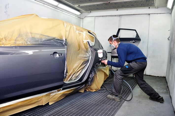 Una persona vernicia a spruzzo un'auto in una cabina di verniciatura. L'auto è parzialmente ricoperta di carta.