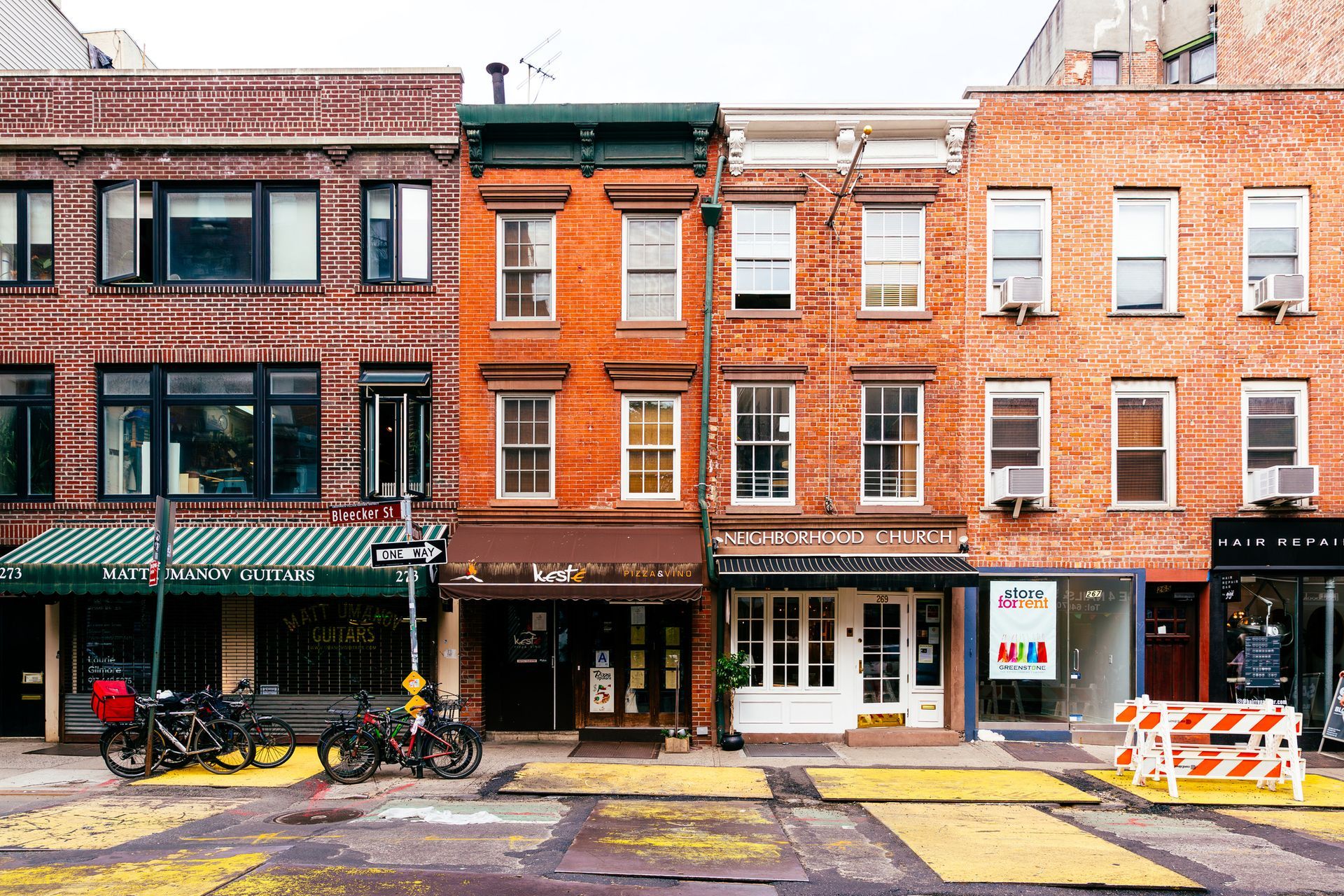 Row of brick buildings with storefronts and a cloudy sky. Yellow crosswalk. Bicycles parked.