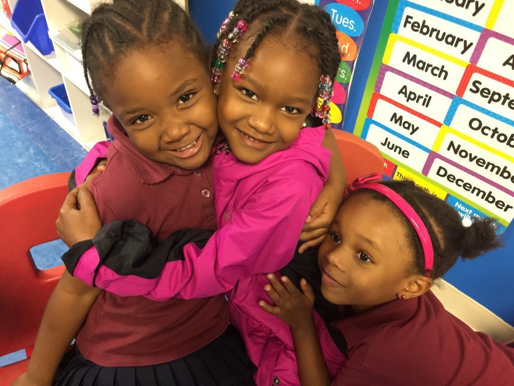 Three little girls are hugging each other in a classroom.