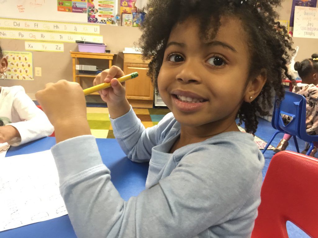 A little girl is sitting at a table with a pencil in her hand.