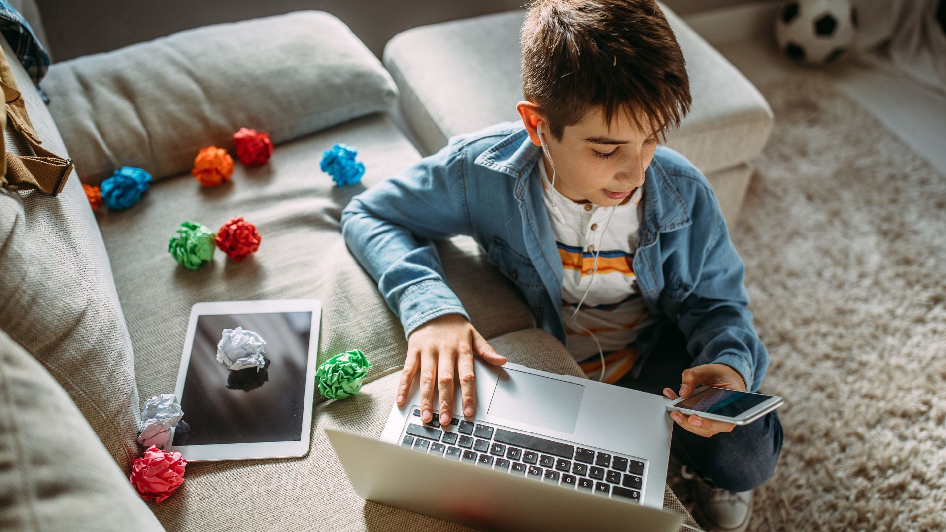 Ein Kind sitzt vor einem Sofa und ist im Netz der digitalen Medien gefangen. Er hat in der einen Hand ein Smartphone und bedient mit der anderen Hand einen Laptop.