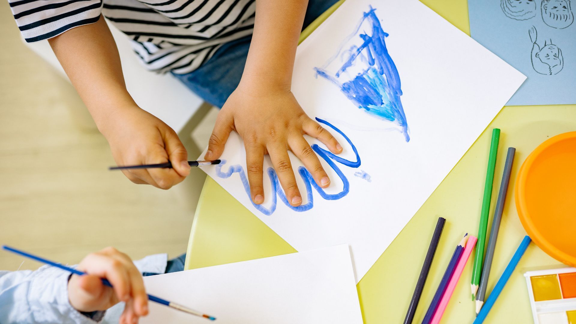 Kinder sitzen an einem Tisch. Sie malen mit Wasserfarbe und Buntstiften blaue Elemente auf das Papier.