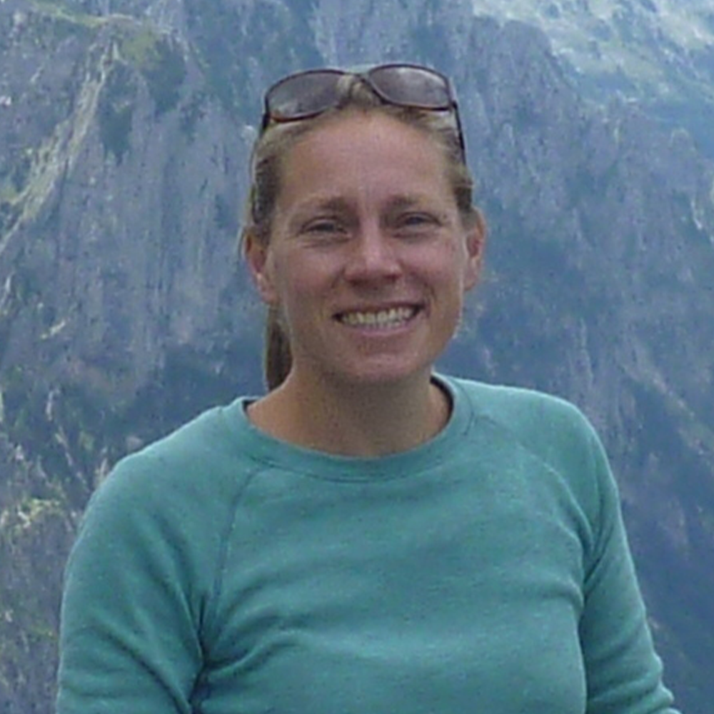 Eine Frau mit Sonnenbrille und blauem Hemd lächelt vor einem Berg.