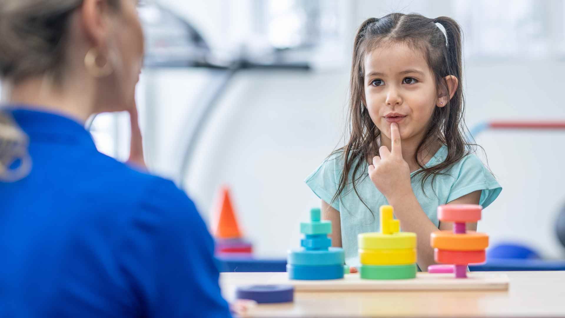 Junges Mädchen am Therapietisch, Finger vor dem Mund, schaut Erwachsenen an. Bunte Spielzeugstapel liegen auf dem Tisch.