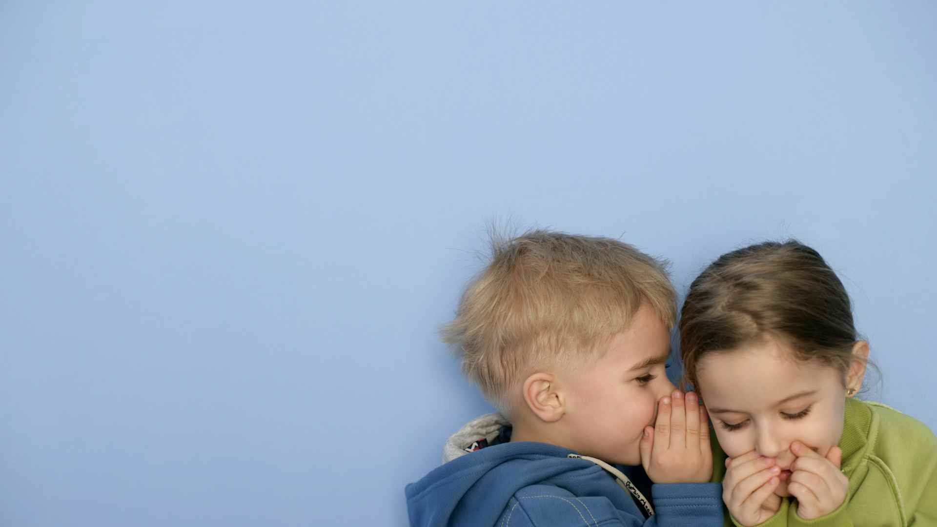 Junge flüstert Mädchen zu, beide lachen. Blauer Hintergrund.
