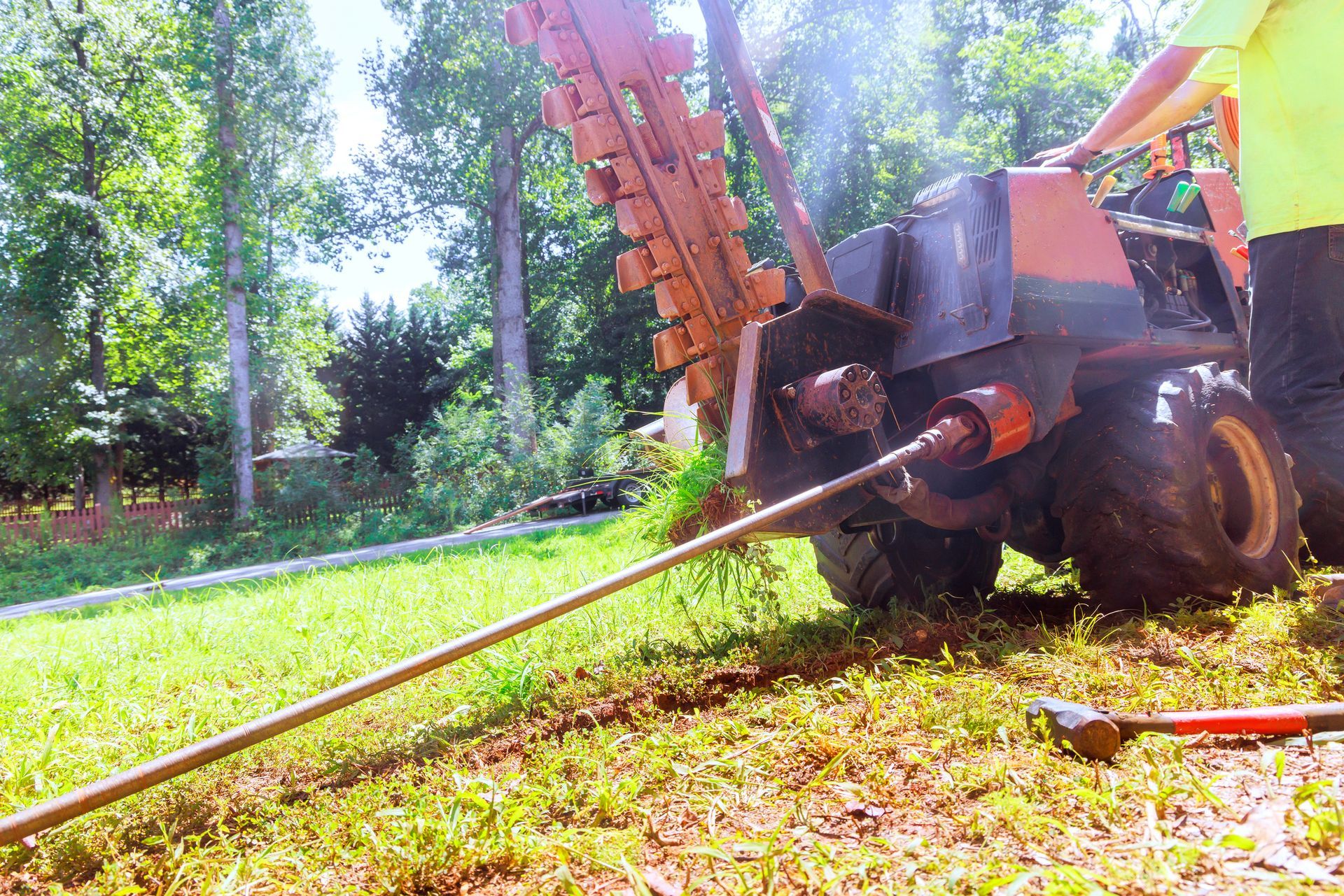 A trencher digging a trench in a grassy area, a person in a safety vest operates the machine.