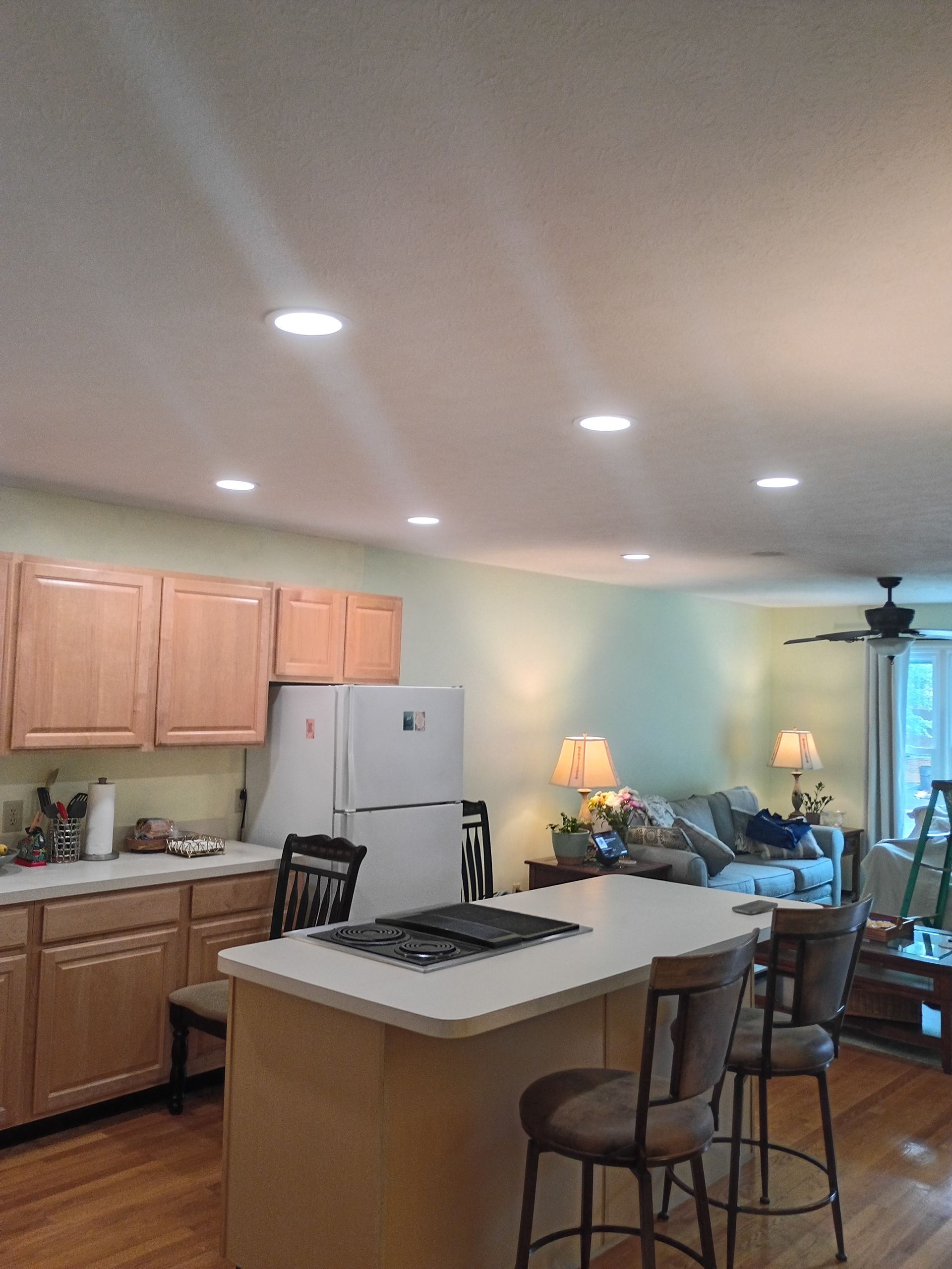 Bright kitchen with light wood cabinets, white island, stools, stove, fridge, and a blue living area beyond.