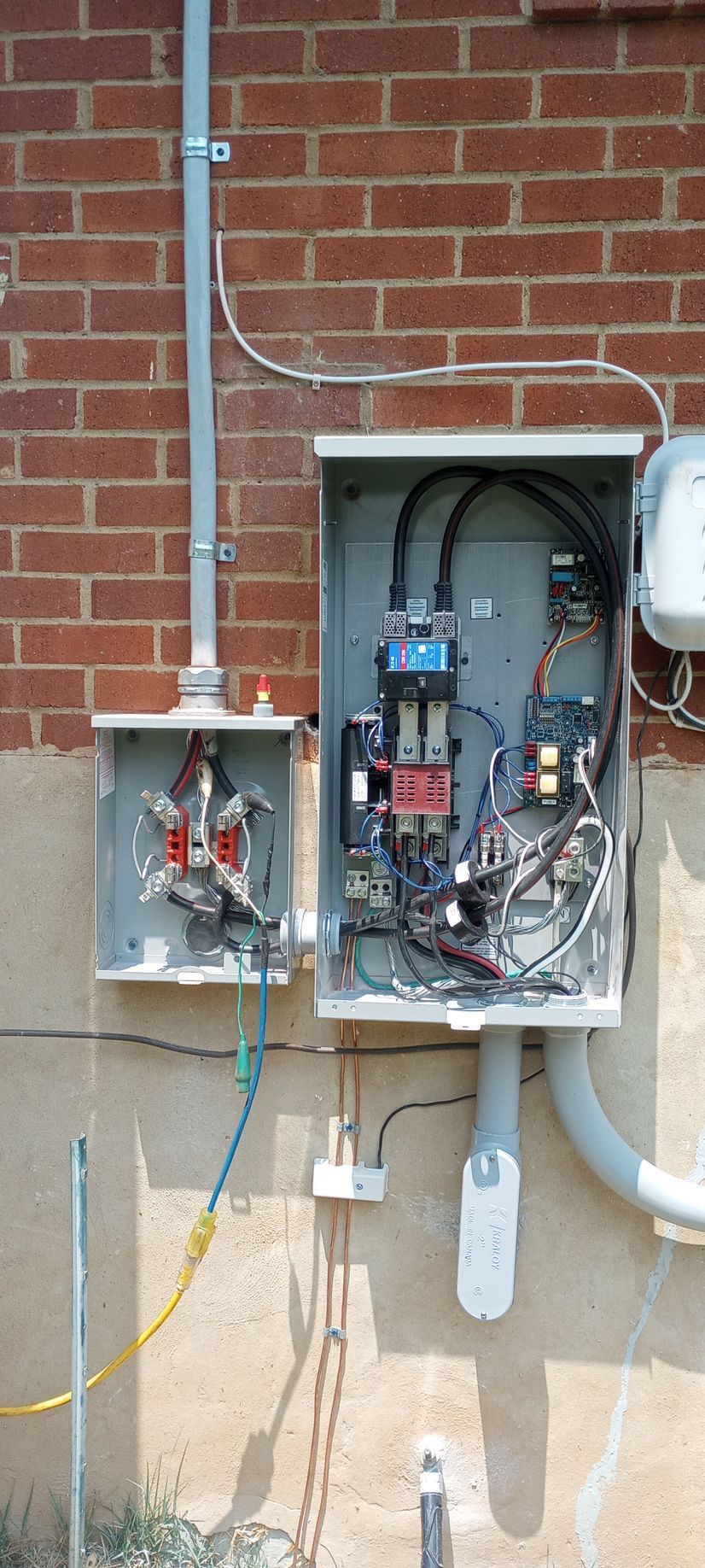 Open electrical service boxes mounted on a brick wall with exposed wiring and pipes.