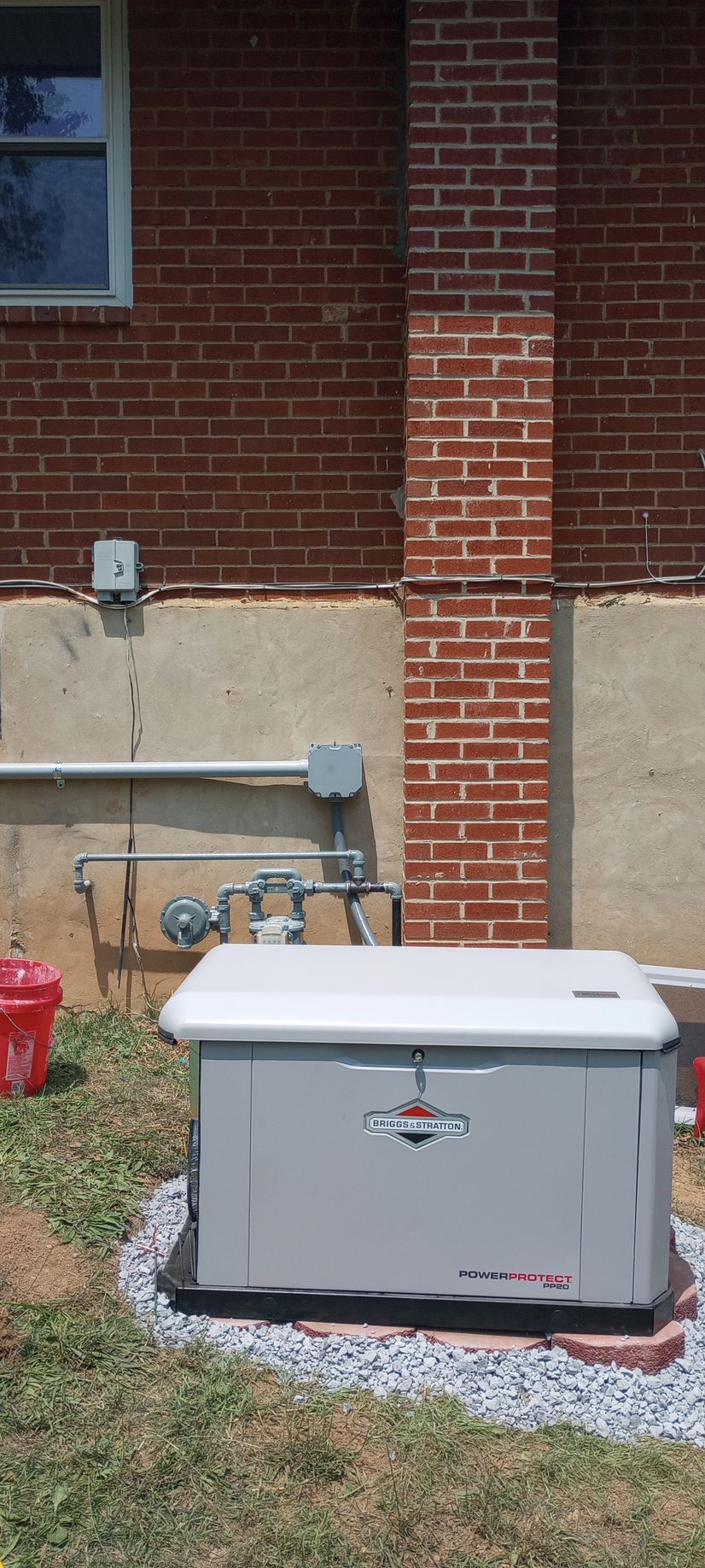 Gray generator on gravel beside a brick house, with utility lines and meters on the wall.