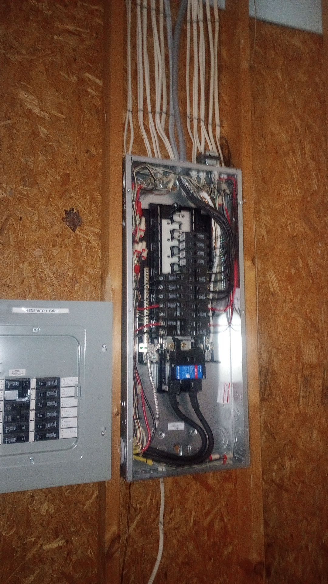 Open electrical breaker panel with wiring inside, mounted on plywood beside another gray panel.