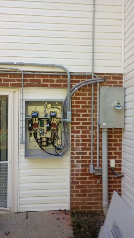 Open electrical panel on exterior brick wall beside gray utility box and conduit piping
