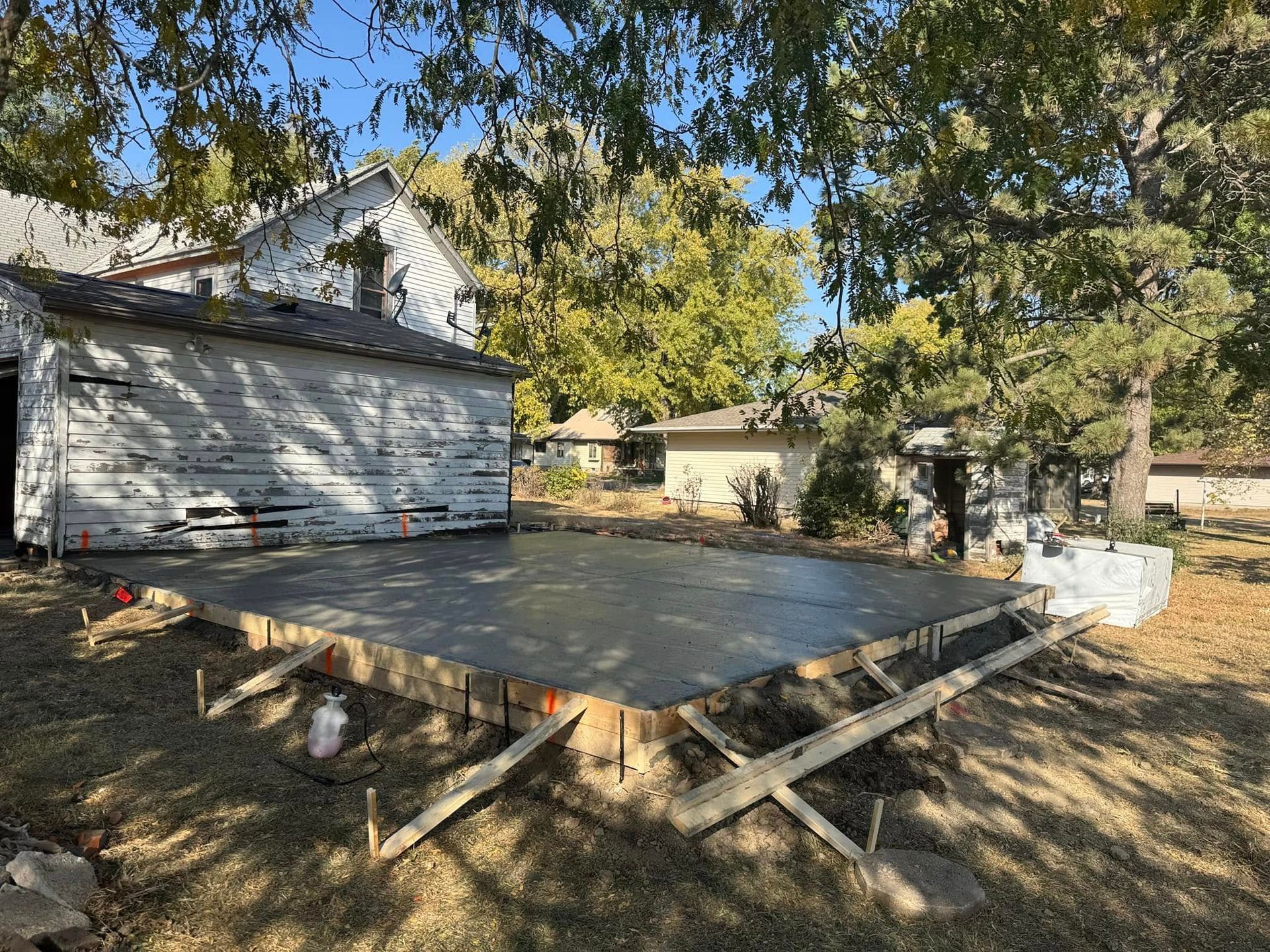 Newly poured concrete slab in a yard, framed by wooden supports, with a weathered building in the background.