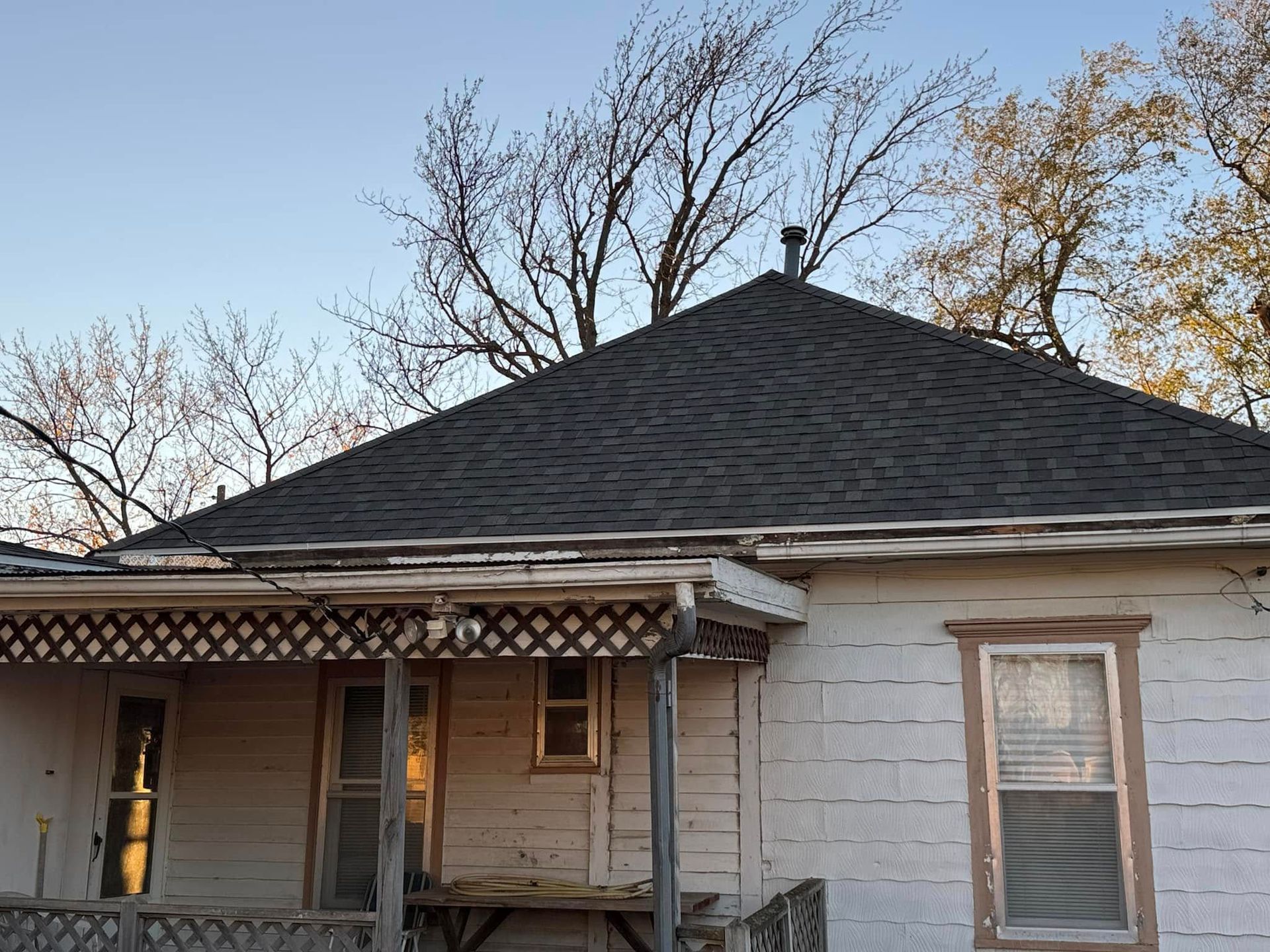 A weathered, white house with a dark shingle roof, surrounded by bare trees under a blue sky.