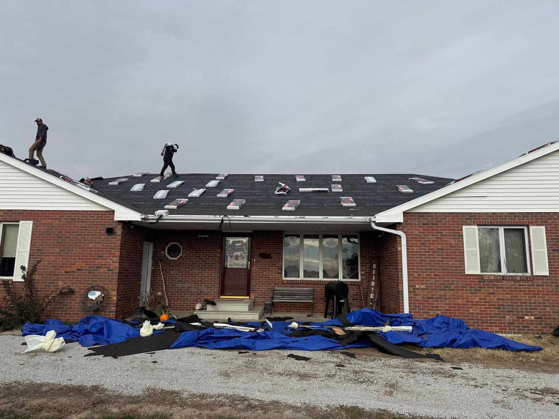 Two people on a damaged roof. Debris and tarp on the ground near a brick house under an overcast sky.