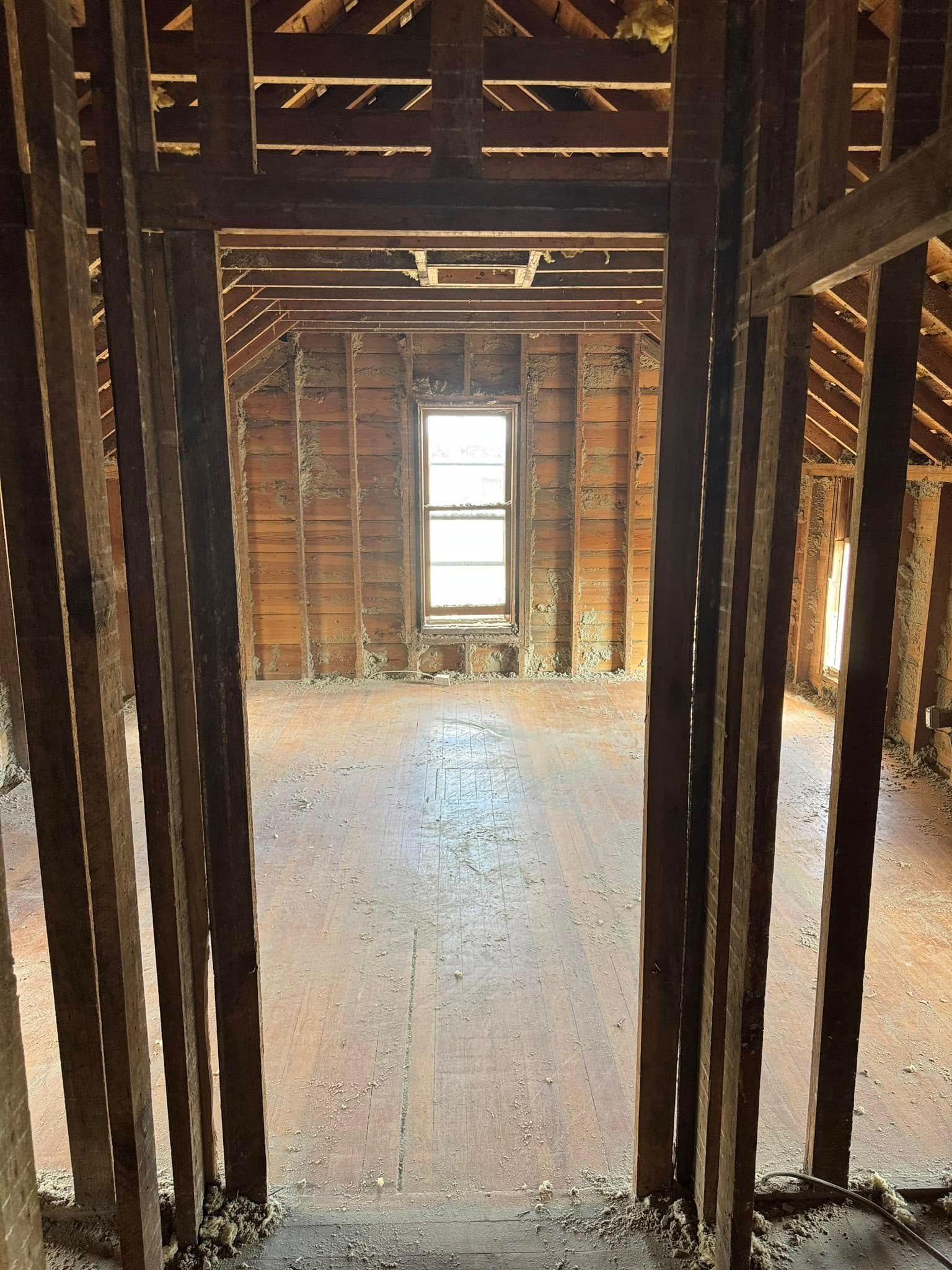Interior of a room undergoing renovation; exposed wooden framing and studs, bare walls, window at the end.