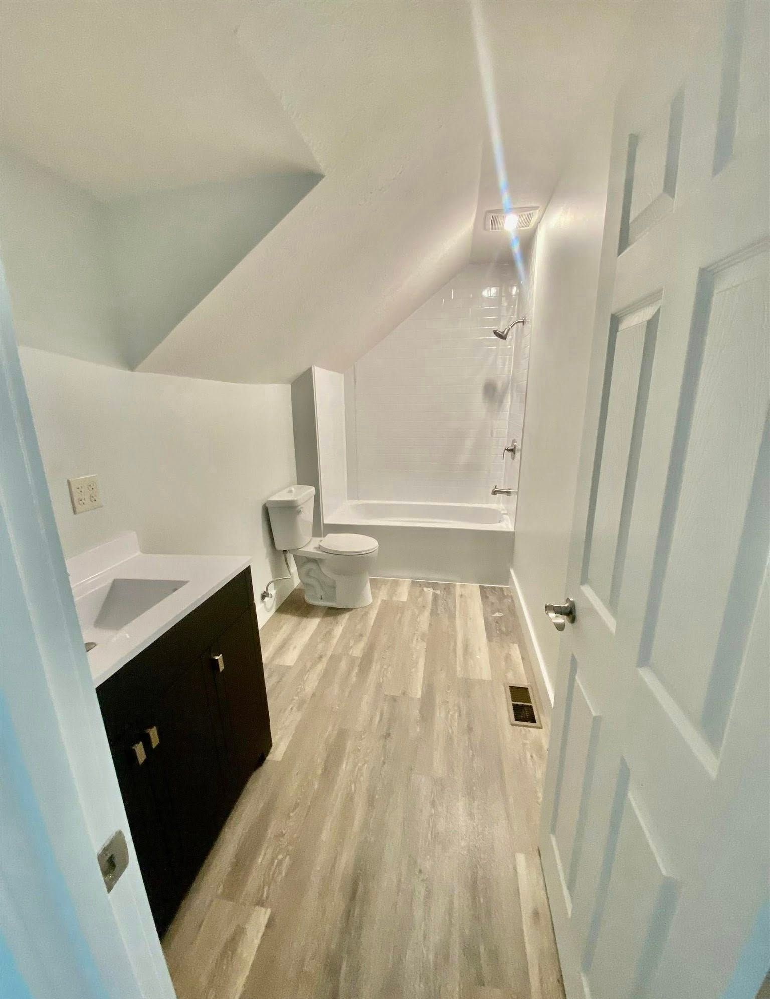 Bathroom with angled ceiling, featuring a vanity, toilet, bathtub, and wood-look flooring.