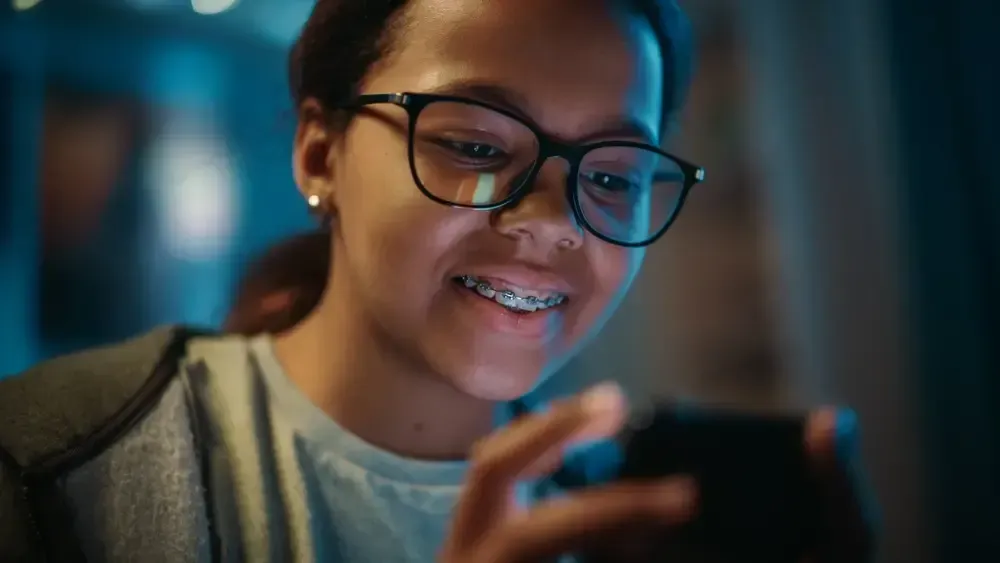 Smiling teenage girl with glasses and braces looking at a smartphone.