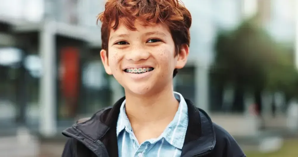 Young boy with freckles and dental braces smiling outdoors.