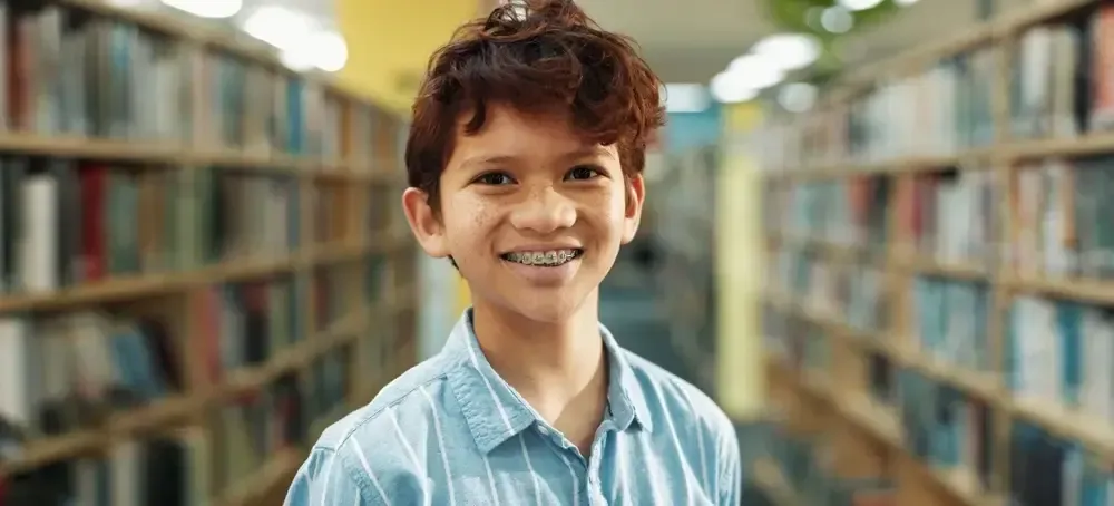 Young boy with braces smiling in a library with bookshelves.