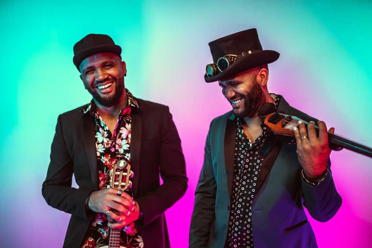 Two men with instruments smiling, vibrant neon background. One with a trumpet, the other with a violin.