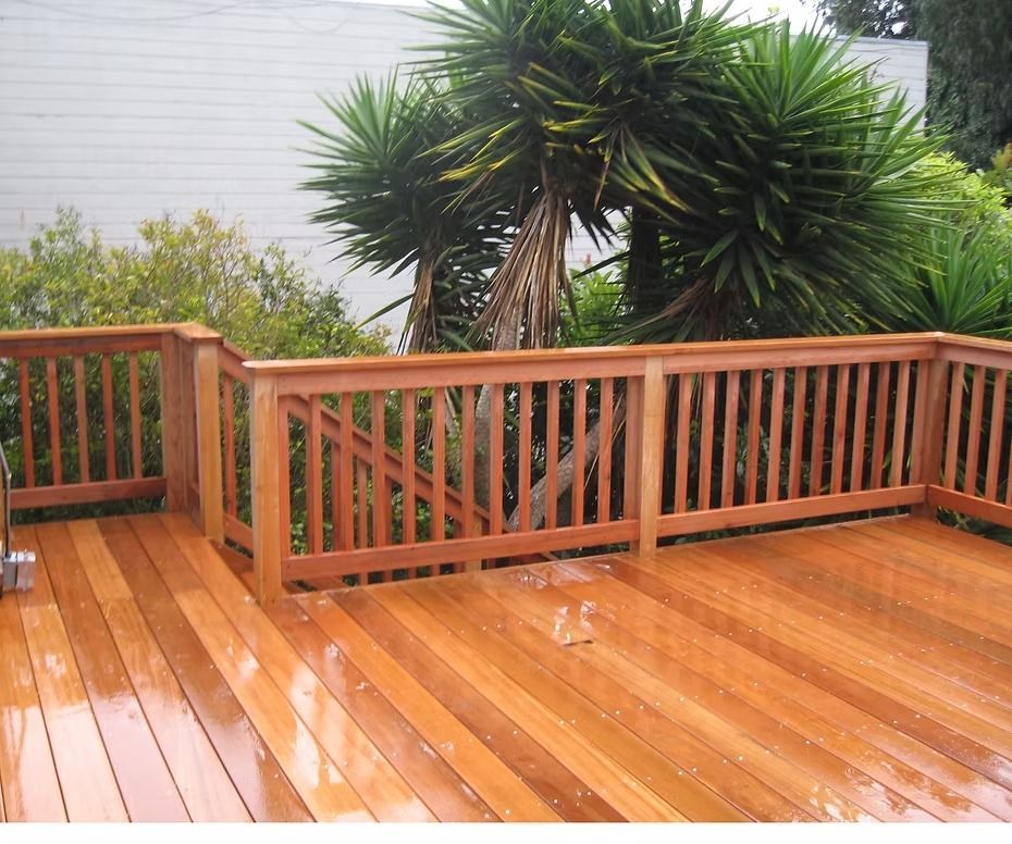 Wooden deck with red-brown railing and siding, white house wall, and a green garden hose on the floor.