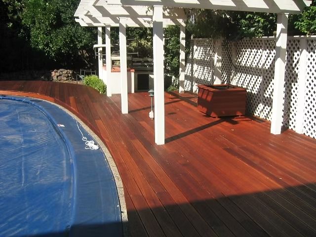 Backyard deck with a blue pool, red-brown wooden floor, white pergola, and a bench in sunlight