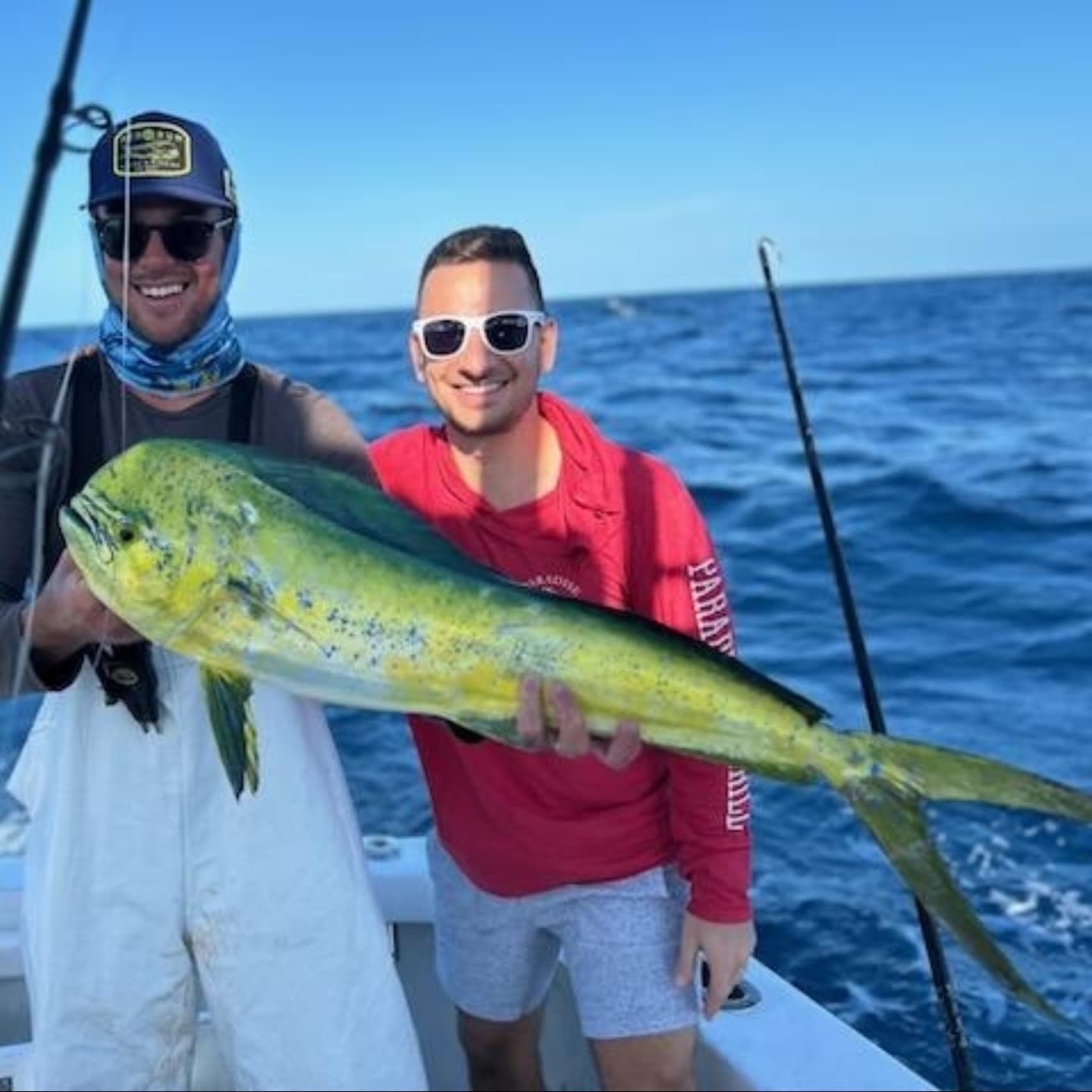 Two people smiling on a boat hold a large, vibrant yellow and green mahi-mahi fish over the ocean.