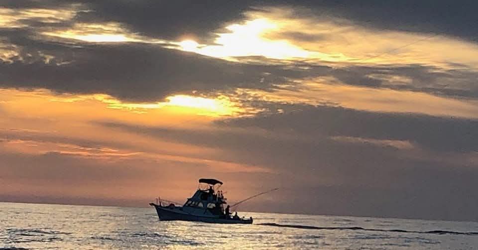 A silhouette of a fishing boat on the open ocean under a cloudy, golden sunset sky.