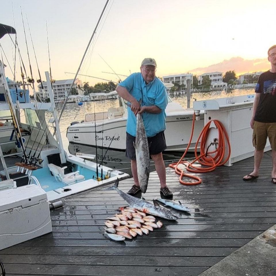 An angler stands on a dock holding a large fish, with a pile of smaller fish and a boat in the background at sunset.