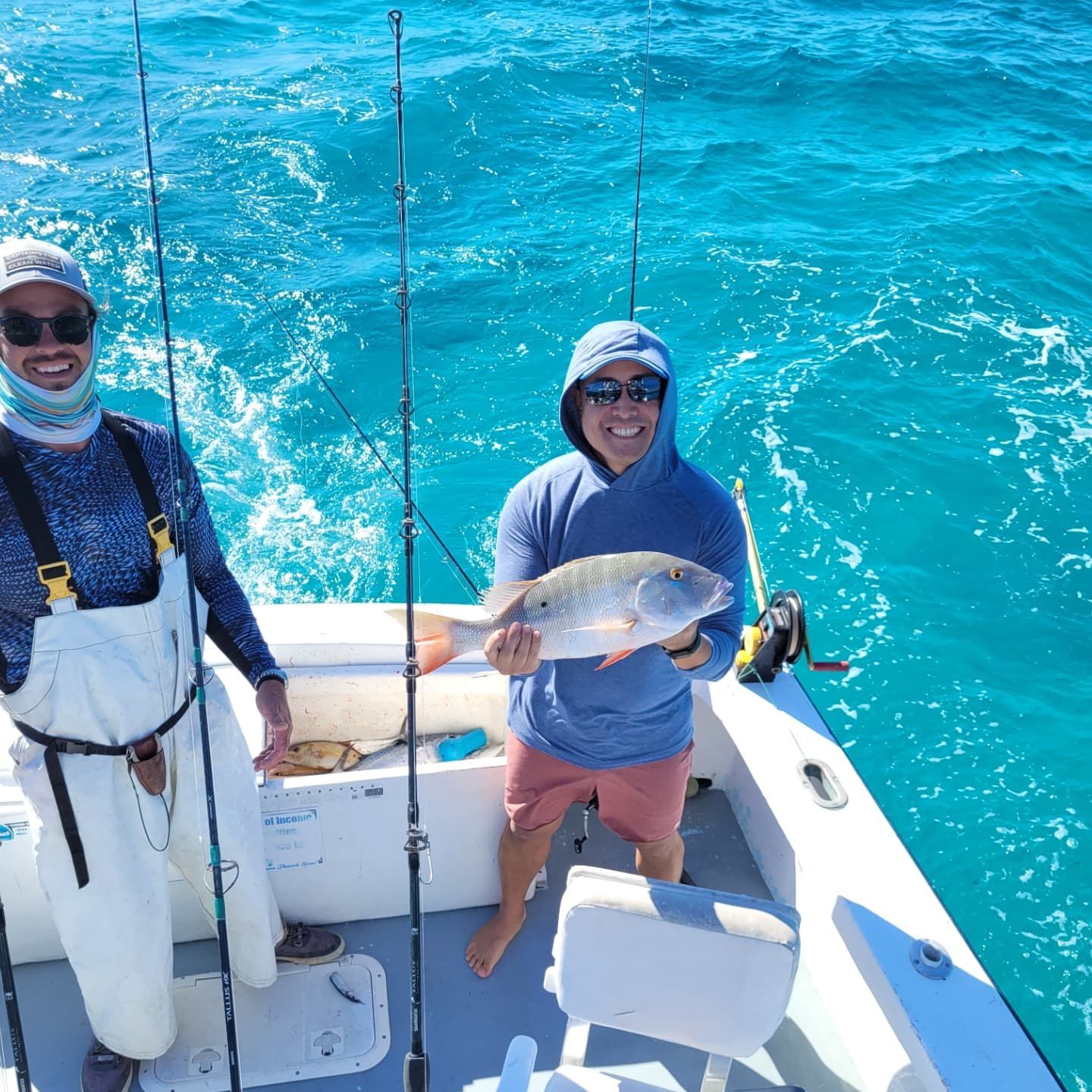 Two people on a boat in bright blue water, one holding a freshly caught fish while the other stands beside them.