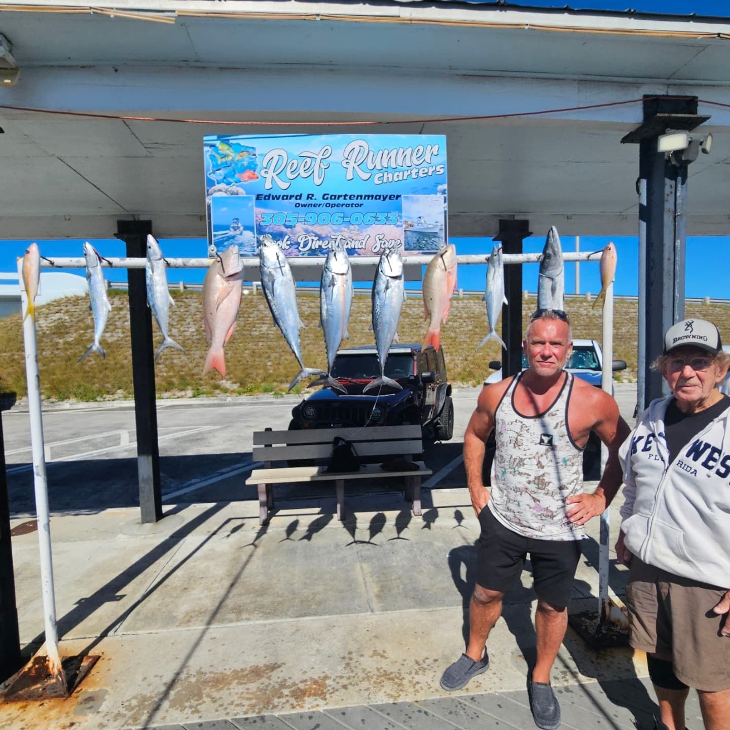 Two people stand under a Reef Runner fishing charter sign with a rack of various fish hanging behind them outdoors.